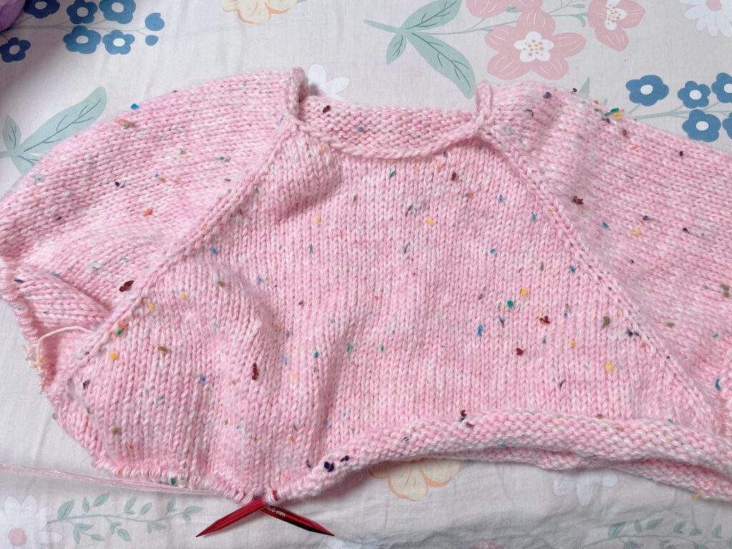 Close-up of a partially knitted pink sweater in progress, showing yarn and stitches. 